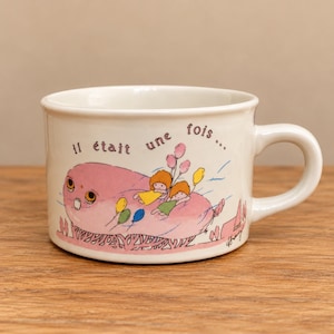 Peut inclure: Un mug en céramique blanche orné d'une illustration fantaisiste représentant une grande créature rose avec deux petits personnages sur son dos tenant des ballons colorés. Le texte "il était une fois..." est imprimé en brun au-dessus.