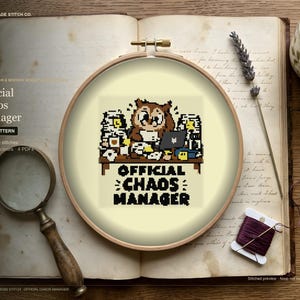 May include: A round wooden embroidery hoop containing a cross-stitch pattern of a brown owl sitting at a desk cluttered with papers and a laptop. The text "OFFICIAL CHAOS MANAGER" is stitched in black pixelated letters below the desk illustration.