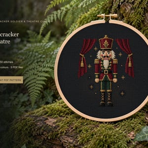May include: A circular wooden embroidery hoop containing a cross-stitch design of a nutcracker soldier standing between two red theatre curtains. The design features gold star accents on a black fabric background. Text reads Nutcracker Theatre.