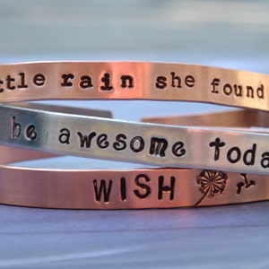 Puede incluir: Tres pulseras de metal apiladas con mensajes inspiradores. La pulsera superior es de cobre y dice "Despu&eacute;s de un poco de lluvia, ella encontr&oacute; su camino." La pulsera del medio es de plata y dice "S&eacute; incre&iacute;ble hoy." La pulsera inferior es de cobre y dice "WISH" con un dise&ntilde;o de diente de le&oacute;n.