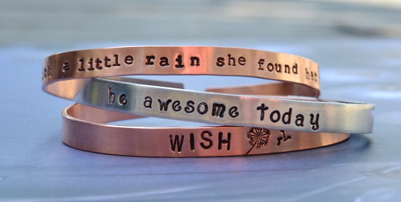 Puede incluir: Tres pulseras de metal apiladas con mensajes inspiradores. La pulsera superior es de cobre y dice "Despu&eacute;s de un poco de lluvia, ella encontr&oacute; su camino." La pulsera del medio es de plata y dice "S&eacute; incre&iacute;ble hoy." La pulsera inferior es de cobre y dice "WISH" con un dise&ntilde;o de diente de le&oacute;n.