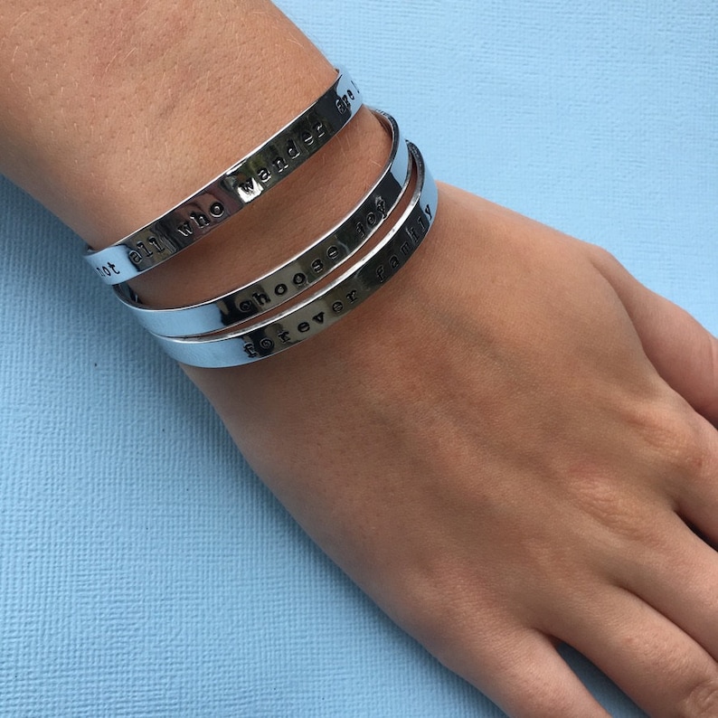 Puede incluir: Tres pulseras de plata apiladas en la mu&ntilde;eca de una persona. Las pulseras tienen el siguiente texto grabado: "Not all who wander are lost", "Choose joy" y "Forever family".
