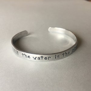 Puede incluir: Una pulsera de metal plateado con la inscripci&oacute;n "All the water in the ocean" grabada en ella.