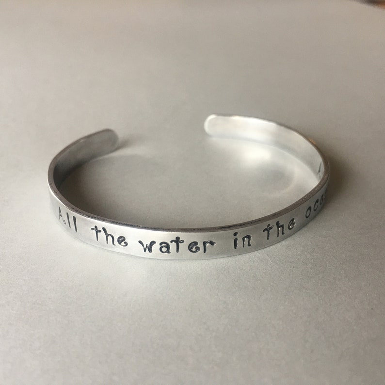 Puede incluir: Una pulsera de metal plateado con la inscripci&oacute;n "All the water in the ocean" grabada en ella.