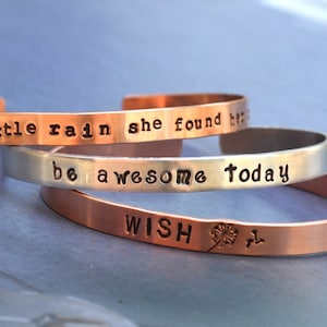 Puede incluir: Tres pulseras apiladas de cobre y plata con mensajes grabados. La pulsera superior dice "with a little rain she found her", la pulsera del medio dice "be awesome today", y la pulsera inferior dice "WISH" con un gr&aacute;fico de diente de le&oacute;n.