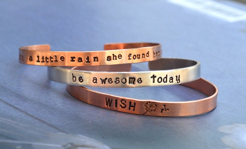 Puede incluir: Tres pulseras apiladas de cobre y plata con mensajes grabados. La pulsera superior dice "with a little rain she found her", la pulsera del medio dice "be awesome today", y la pulsera inferior dice "WISH" con un gr&aacute;fico de diente de le&oacute;n.