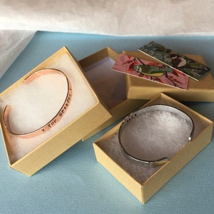 Puede incluir: Dos peque&ntilde;as cajas con tapas, una que contiene un brazalete de cobre grabado con el texto "x for Grandma", la otra que contiene un brazalete de plata grabado con el texto "Miss".