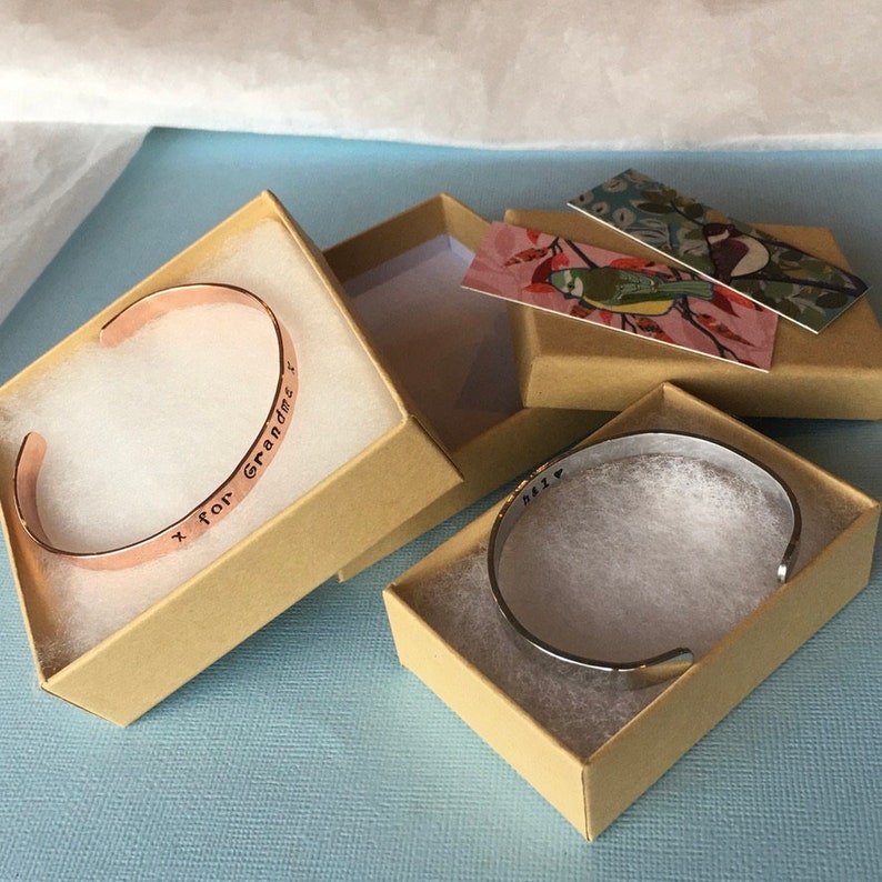 Puede incluir: Dos peque&ntilde;as cajas con tapas, una que contiene un brazalete de cobre grabado con el texto "x for Grandma", la otra que contiene un brazalete de plata grabado con el texto "Miss".