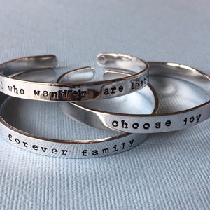 Puede incluir: Tres pulseras de plata con grabados que dicen: "forever family", "who wander are lost" y "choose joy".