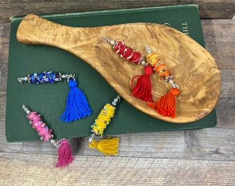 May include: Five colorful tassel keychains with beaded accents. The keychains are arranged on a wooden surface and a green book with the word "THE" visible on the cover.