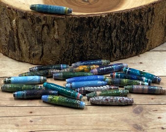 May include: A collection of colorful, cylindrical beads in various shades of blue, green, and patterned designs. The beads are scattered on a wooden surface, with a piece of wood in the background.