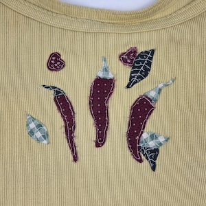 May include: A pale yellow ribbed tank top featuring fabric appliqué patches of three dark red chili peppers, two small round berry-like shapes, and four leaves in green and white checkered or dark patterned fabric stitched onto the front.