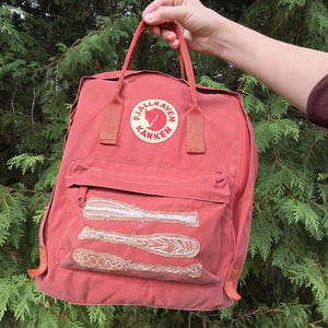 May include: A terracotta-colored Fjällräven Kånken backpack featuring two top handles, a front zippered pocket, and three embroidered baseball bat designs on the front pocket. The circular logo patch displays the text Fjällräven Kånken.