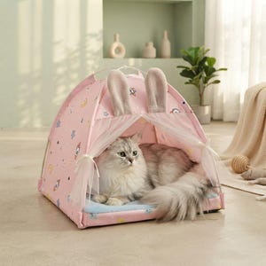 Puede incluir: Una tienda de campaña rosa para mascotas con un estampado de pequeños cohetes, estrellas y lunas. La tienda tiene dos orejas de conejo de felpa erguidas en la parte superior y cortinas blancas transparentes atadas en la entrada. Un gato de pelo largo descansa dentro sobre una alfombrilla azul suave.