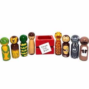 May include: A set of eight hand-painted wooden peg dolls representing various animals, including a frog, lion, snake, and monkey, arranged around a small red wooden crate with a white tag that reads FROM THE ZOO in black capital letters.