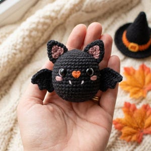 Peut inclure: Une petite chauve-souris en crochet noire, ronde, avec deux yeux en plastique noir, un petit nez en forme de cœur orange, deux crocs blancs et des joues brodées en rose. Elle possède deux ailes noires et des oreilles avec des détails rose clair.
