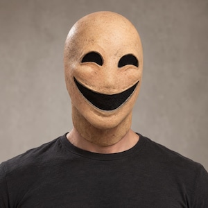 Puede incluir: Una máscara de cabeza completa con un acabado texturizado de color beige similar a la piel, que presenta una sonrisa amplia, exagerada y sin dientes, junto con aberturas para los ojos en forma de almendra y malla negra en el interior.