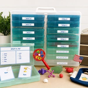 May include: A teal and white plastic storage case with labeled compartments for phonics lessons. The case contains flashcards, toys, and an American flag. The labels read "short a", "long a", and other vowel sounds.