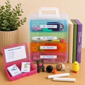 May include: A clear plastic organizer with four colored compartments labeled with digraphs. The organizer contains small objects like a toy shark, a pencil, and a small chair. A pink case with digraph flashcards and a small wooden chair are in front.