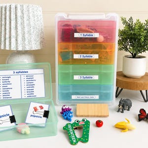May include: A colorful educational set for learning syllables. It includes a multi-tiered storage container with labels for 1, 2, and 3 syllables, along with word and picture cards. Additional toy figures and a lamp are also present.