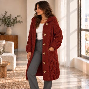 May include: A long-sleeved, knee-length, dark red cable-knit cardigan with a hood and large light-coloured buttons down the front. The garment features thick, textured cable patterns on the sleeves and front pockets, worn over a white top and grey trousers.