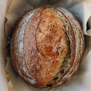 May include: A round loaf of sourdough bread with a dark, golden-brown crust and a prominent score line down the center. The bread is resting inside a round cooking vessel lined with light tan parchment paper, showing a textured, baked surface.
