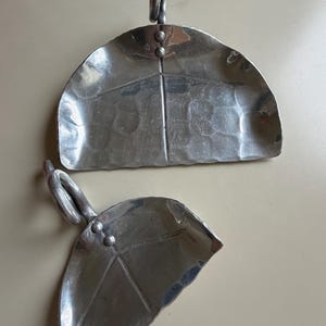May include: Two silver-toned metal decorative scoops with a hammered texture. Each scoop features a semi-circular bowl, a central line detail, two small circular accents near the top, and a curved handle loop for hanging or display purposes.