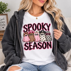 May include: A white t-shirt featuring the text SPOOKY SEASON in black and pink brushstroke lettering. Below the text are five ghost illustrations wearing various accessories including a leopard print bow, a pink cowboy hat, a bow, a black cowboy hat, and heart-shaped sunglasses, layered over a dark gray zip-up hoodie.