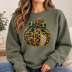 Peut inclure: Un sweat-shirt à col rond vert olive avec un motif central de citrouille. La citrouille présente un imprimé camouflage, un nœud vert foncé et une section à motifs de feuilles blanches et vertes. Le dessin est souligné d'un contour doré.