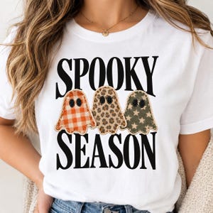 Może przedstawiać: Biały t-shirt z czarnym napisem SPOOKY SEASON i trzema ilustracjami duchów. Lewy duch ma pomarańczowy wzór w kratkę, środkowy brązowy wzór w panterkę, a prawy zielony wzór w gwiazdki.