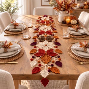 May include: A crochet table runner featuring a repeating pattern of autumn leaves in shades of cream, orange, red, brown, and deep purple. The runner is decorated with small crocheted berries and is centred on a wooden dining table with place settings.