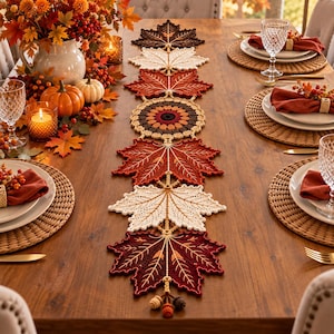 May include: A crochet table runner featuring a series of interconnected maple leaf motifs in shades of dark brown, cream, orange, and deep red. The runner includes a central circular mandala pattern and ends with small dangling acorn-shaped embellishments.