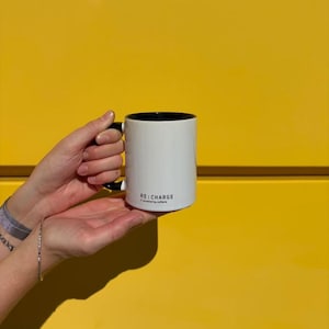Op de afbeelding: Een witte keramische mok met een zwarte binnenkant en een zwart handvat. De buitenkant is voorzien van de zwarte tekst RE:CHARGE // powered by caffeine. De mok heeft een cilindrische vorm en een gladde afwerking voor warme dranken.