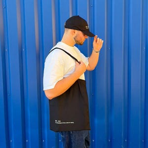 Może przedstawiać: Czarna czapka z daszkiem z białym napisem, gładki biały t-shirt z krótkim rękawem oraz czarna torba typu tote z nadrukowanym białym tekstem RE:DO i frazą pretending to know what I'm doing, zestawione z ciemnoniebieskimi dżinsami.