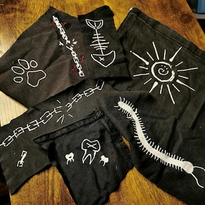 Könnte beinhalten: Eine Sammlung schwarzer Stoffaufnäher mit handgemalten weißen Motiven, darunter ein Pfotenabdruck, ein Fischskelett, eine Sonne, ein Tausendfüßler, ein Zahn und Kettenglieder. Die Aufnäher haben rohe, unebene Kanten und sind verstreut angeordnet.