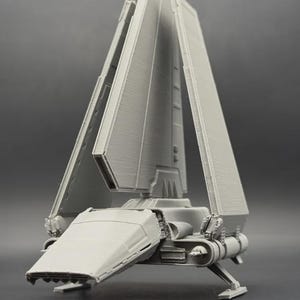Peut inclure: Un modèle imprimé en 3D gris clair représentant une navette spatiale de classe Lambda. Le véhicule présente une dérive verticale centrale proéminente, deux grandes ailes repliables orientées vers le haut, un cockpit effilé et des trains d'atterrissage.