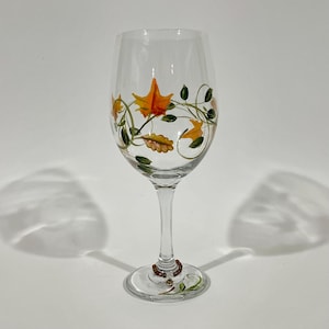 May include: A clear glass wine glass featuring hand-painted orange and yellow autumn leaves with green vines and small dark berries. The stem of the glass is adorned with a small beaded charm near the base, which also has painted vines.