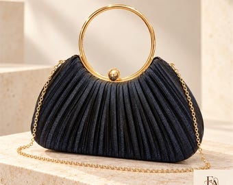 May include: A dark navy blue pleated clutch bag with a shimmering texture, featuring a circular gold metal handle and a matching gold chain shoulder strap. The bag is secured with a small gold ball clasp. The logo Freya Atelier is visible.