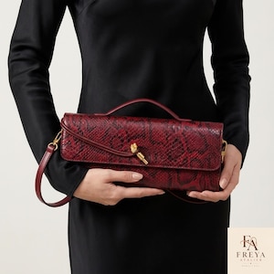 May include: A rectangular handbag featuring a deep red and black snakeskin pattern. The bag has a top handle, a detachable shoulder strap, and a gold-toned metal knot detail on the front flap. The brand name Freya Atelier is printed in the corner.