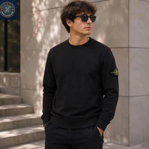Puede incluir: Una sudadera negra de manga larga con cuello redondo que presenta un parche circular amarillo y verde estilo brújula en la manga izquierda. La prenda tiene cuello, puños y dobladillo acanalados, ofreciendo un aspecto sólido y limpio.