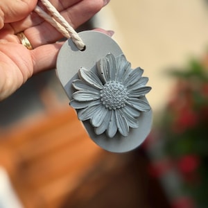 May include: A light gray concrete ornament featuring a raised daisy flower relief on an oval-shaped base. The piece includes a hole at the top with a thick, off-white twisted cotton cord threaded through for hanging purposes.