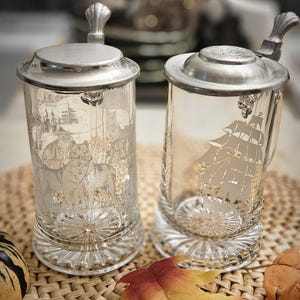 May include: Two clear glass beer steins with metal lids and handles. The left stein features an etched scene of wolves in a forest. The right stein displays an etched sailing ship. Both have a starburst pattern on the base and metal thumb levers.