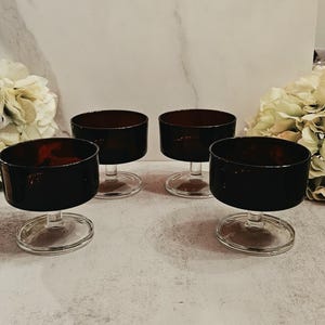 May include: A set of four matching dessert bowls made of dark amber glass. Each bowl features a wide, cylindrical basin supported by a short, clear glass stem and a circular base, providing a stable foundation for serving.