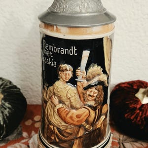 May include: A ceramic beer stein with a conical metal lid featuring an embossed scene of Rembrandt with Saskia. The dark background highlights the figures in period clothing, with the text Rembrandt met Saskia printed in white on the side.