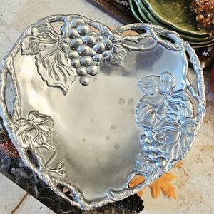 May include: A heart-shaped silver-toned metal serving tray featuring embossed grape clusters and vine leaf motifs along the edges. The smooth, reflective center surface is framed by a detailed border of sculpted metal foliage and fruit.
