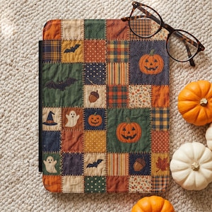 Op de afbeelding: Een rechthoekige e-readerhoes met een patchworkontwerp met diverse herfst- en Halloween-motieven zoals pompoenen, spoken, vleermuizen, eikels en bladeren in oranje, groene, blauwe en bruine tinten, gecombineerd met een bril met donker montuur.
