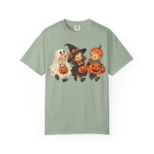 May include: A sage green t-shirt featuring a graphic illustration of three children in Halloween costumes. One child is dressed as a ghost, one as a witch, and one as a pumpkin. Each child is holding a small orange jack-o'-lantern candy bucket.