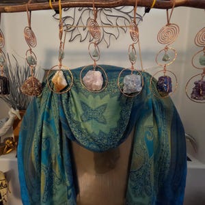 May include: Six hanging ornaments featuring copper wire spirals and circular frames, each holding a raw gemstone. The stones include black, brown, yellow, pink, light blue, dark blue, and purple crystals, suspended from a wooden branch.