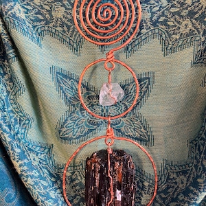 May include: A vertical wall hanging featuring a copper wire spiral at the top, a middle copper circle containing a clear raw crystal, and a bottom copper circle holding a large, dark, vertically striated raw mineral specimen.