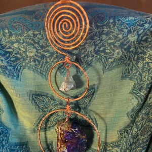 May include: A hanging ornament featuring a hammered copper wire spiral at the top, followed by two circular copper wire frames. The middle frame holds a small, pale green crystal, and the bottom frame holds a large, iridescent purple and gold mineral.